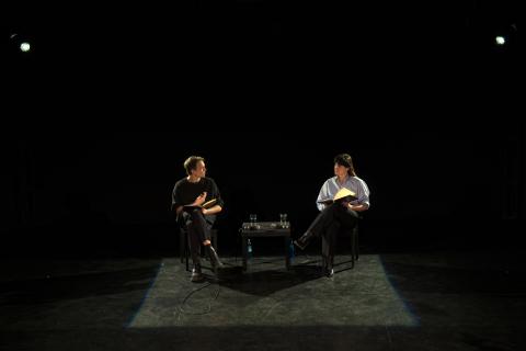 Ein Schauspieler und eine Schauspielerin sitzen in einem dunklen Raum auf Stühlen und sprechen zueinander 