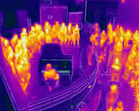 Thermografie-Aufnahme eines Konzerts im Foyer der HfMDK mit Menschen, die um einen Flügel herumstehen.