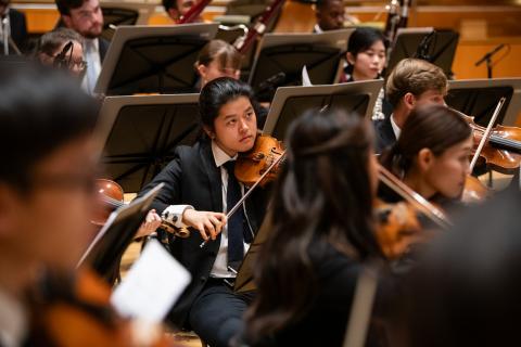 Ein Student inmitten des Orchesters spielt Geige und schaut zum Dirigenten