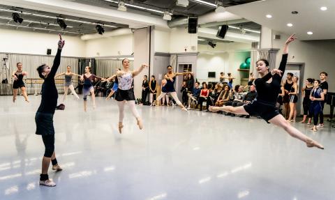 Tänzer:innen mitten im Sprung (grand jeté), die Ballettlehrerin zeigt nach oben, hinter den Studierenden sitzen Zuschauer:innen.