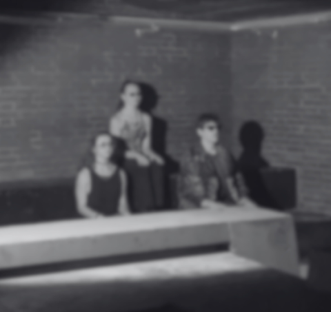 Das schwraz-weiß Foto zeigt in Unschärfte drei Personen sitzend vor einer Wand.