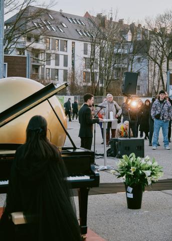 Ein junger Mann in schwarzem Anzug hält eine Rede. Davor sieht man die goldene Kugel und die Pianistin.
