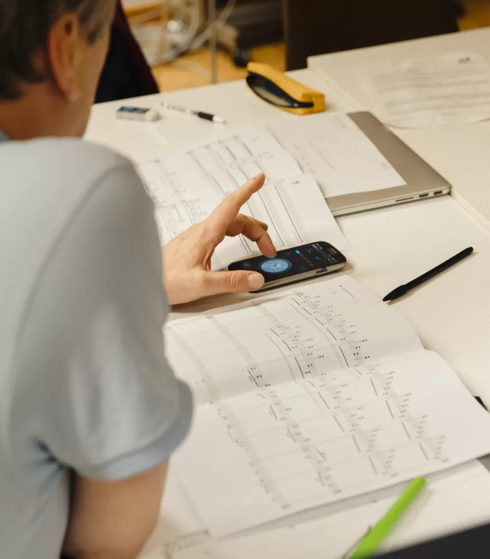 Nahaufnahme einer Hand, die mit dem Finger auf einen Handybildschirm tippt. Daneben Noten auf Papier.