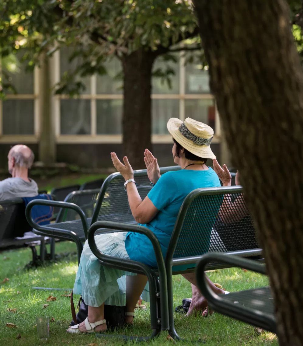 Eine Person mit Strohhut sitzt auf einer Bank und applaudiert.