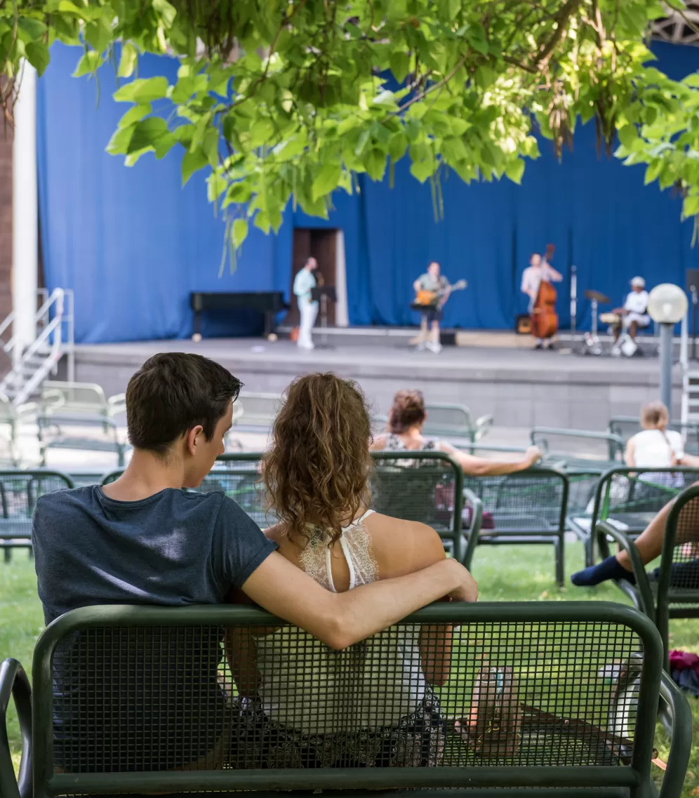 Eine junge Frau sitzt im Arm eines jungen Mannes auf einer Bank. Auf der Bühne spielt ein Jazzquartett