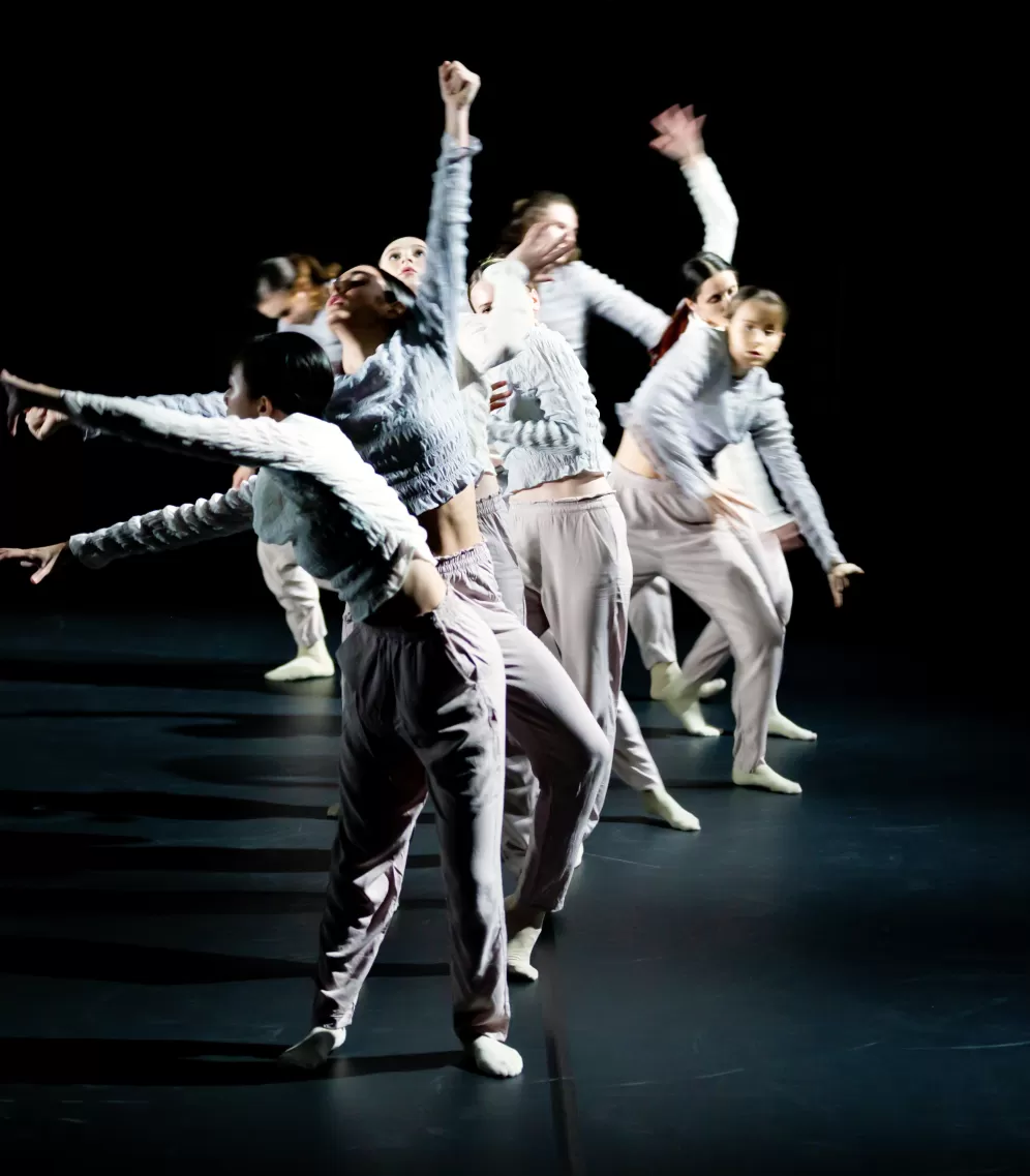 Eine Gruppe Tänzerinnen in Performance, hell gekleidet vor dunklem Bühnenhintergrund. 