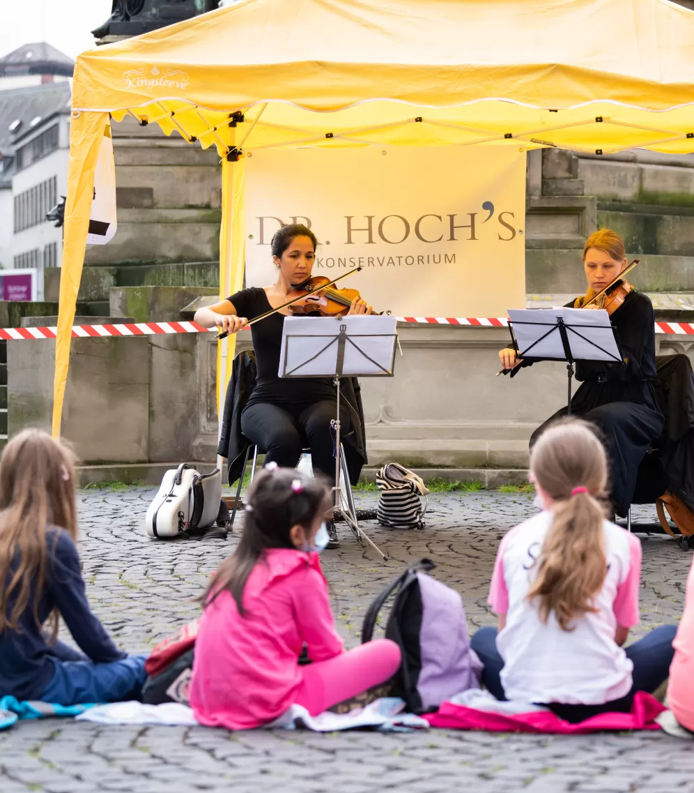 Zwei Musikerinnnen spielen unter einem gelben Pavillon, davor auf dem Boden sitzen mehrere Kinder.