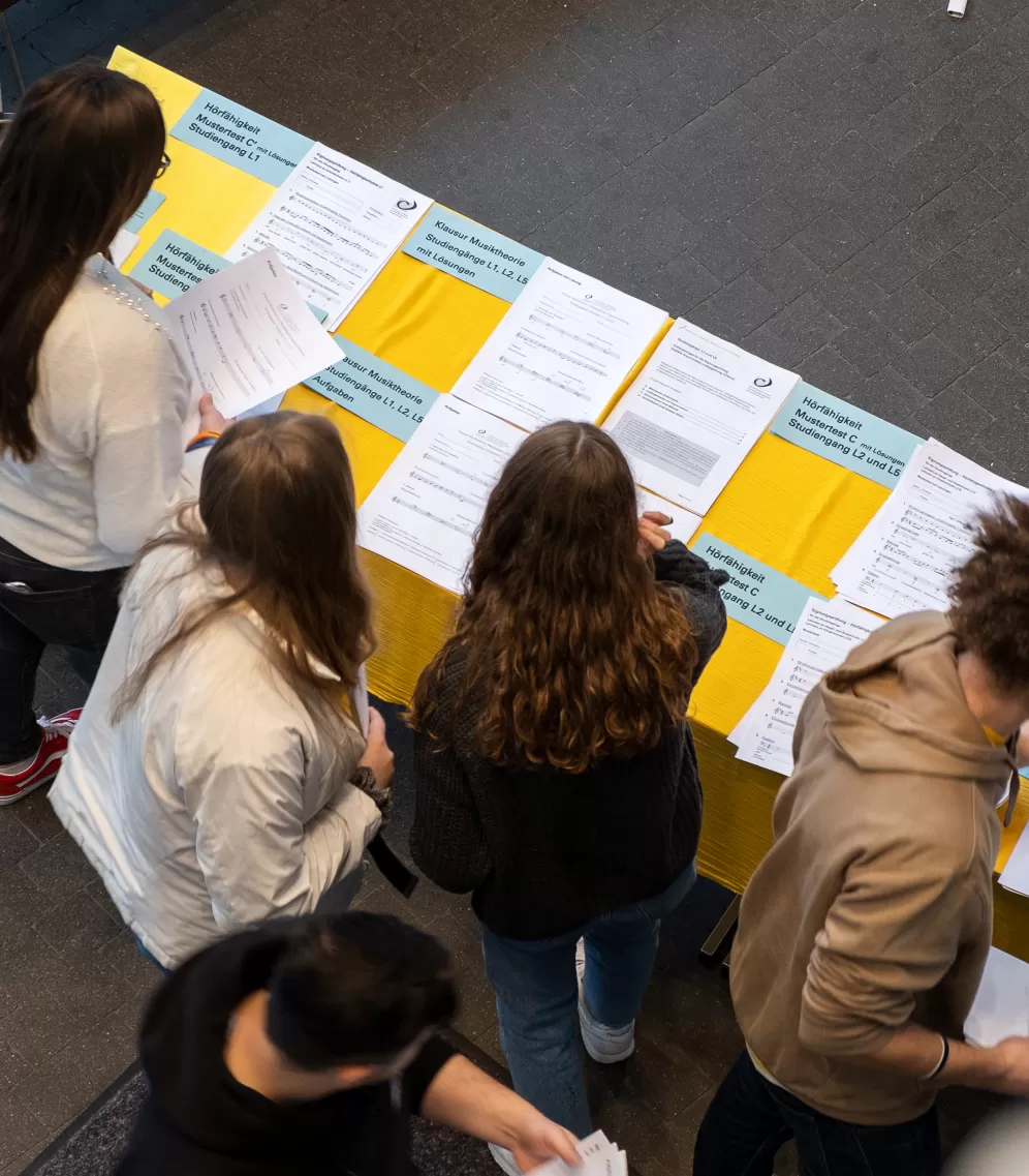 Mehrere Personen stehen an einem Tisch mit Beispieltests für die Eignungsprüfung