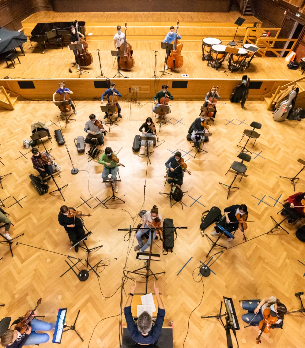 Blick aus der Vogelperspektive auf das Hochschulorchester, das mit Abstand im gesamten Großen Saal verteilt sitzt. Auf dem Boden kleben schwarze Markierungen.