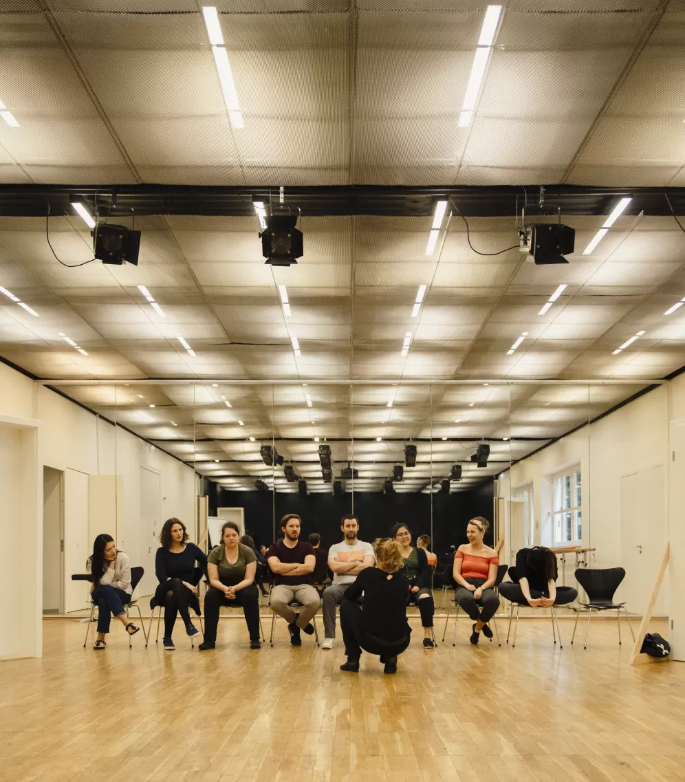 Acht Gesangsstudierende sitzen nebeneinander in einer Stuhlreihe im Opernstudio vor ihnen hockt eine Person