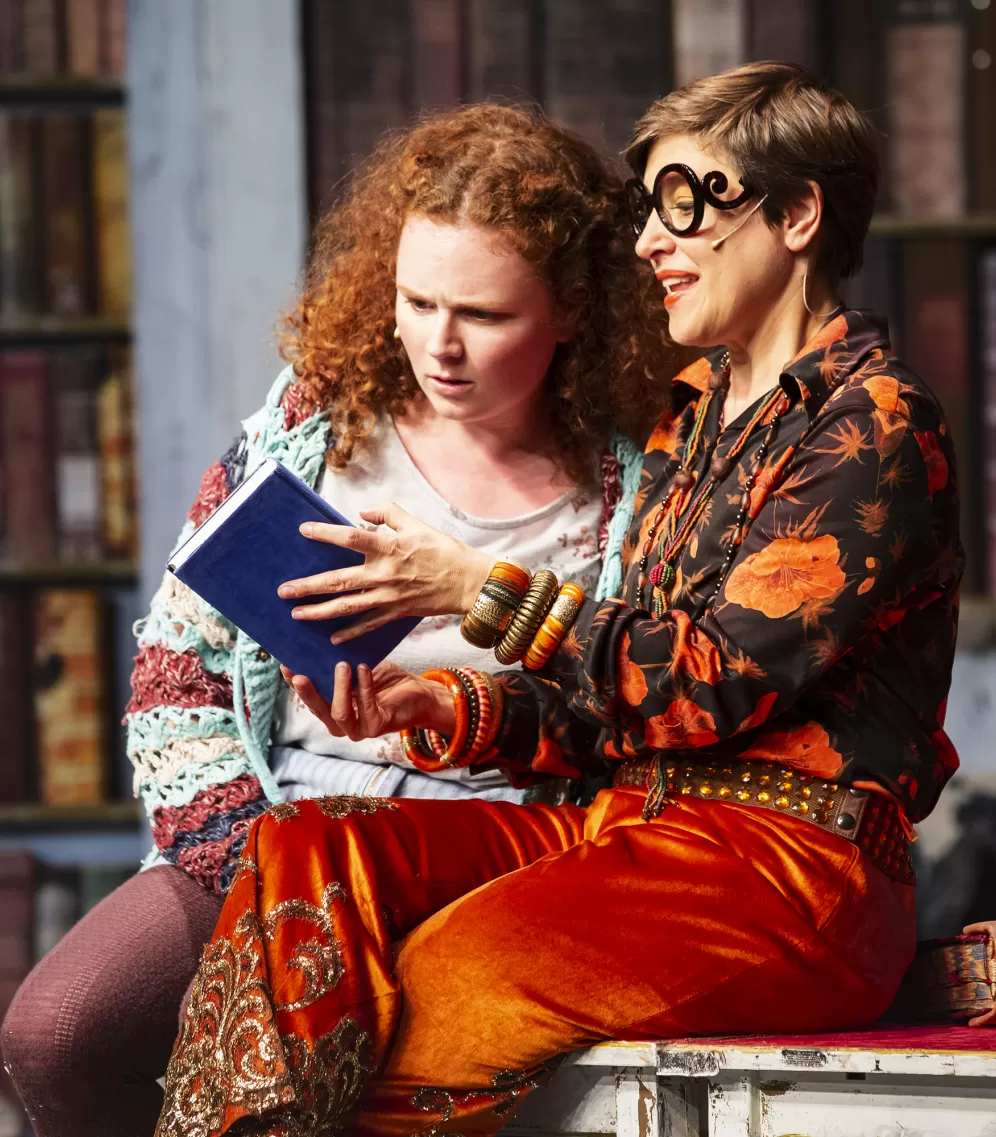 Zwei Schauspielerinnen sitzen auf einem Tisch vor großen Bücherregalen und schauen sich ein blaues Buch an.