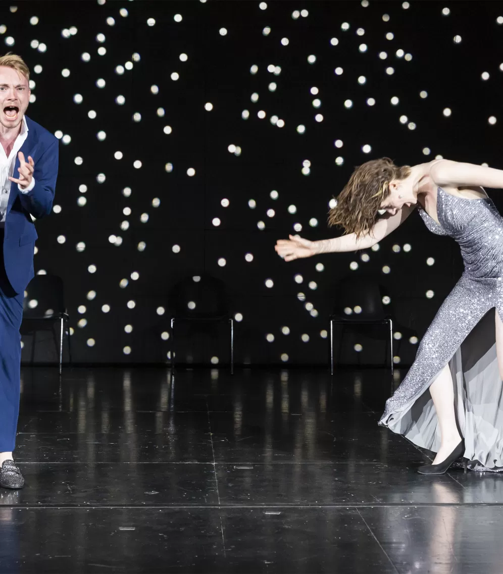 Schauspieler in blauem Anzug, dynamische Bewegung, und Schauspielerin in silbernem Glitzerkleid, seitlich und in Bewegung.
