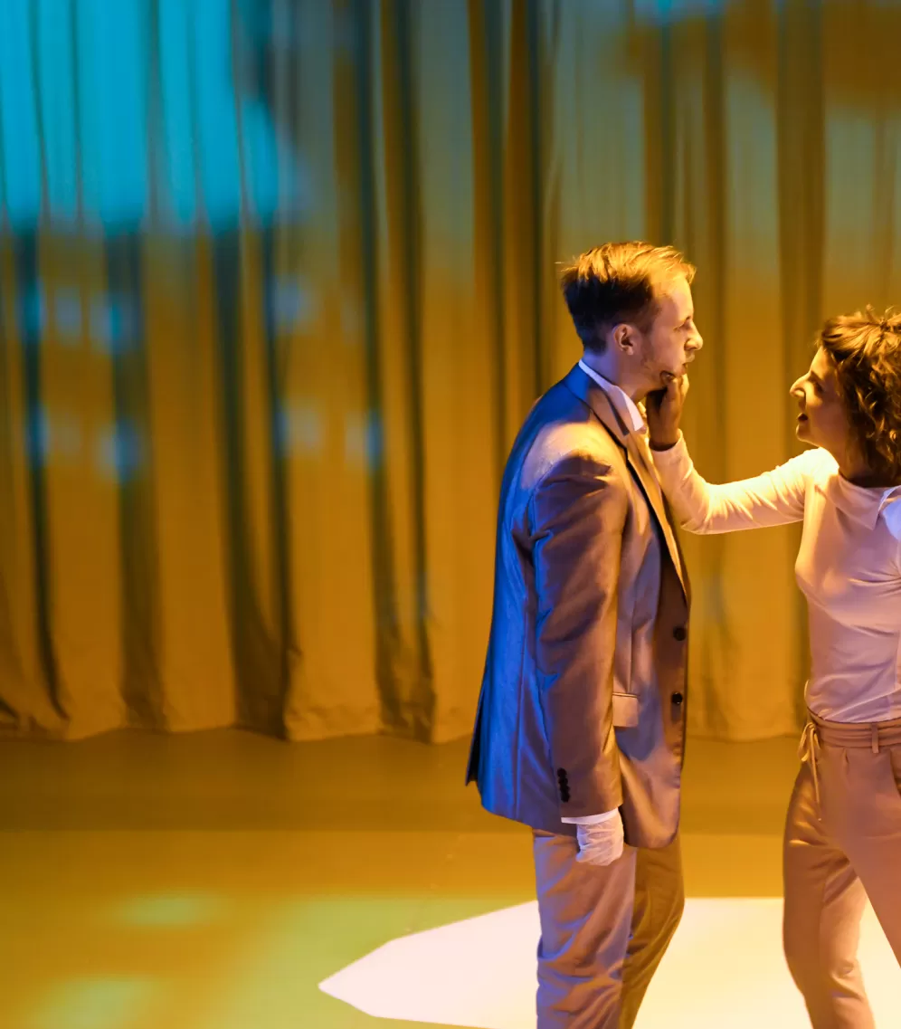 Schauspielerin greift das Gesicht ihres Gegenüber. Leere Bühne, warmes Licht.