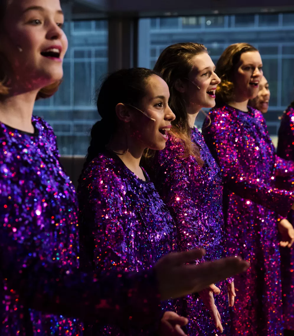 Ensemble mit lila Glitzerkleidern singen mit nach vorne ausgestreckten Händen.