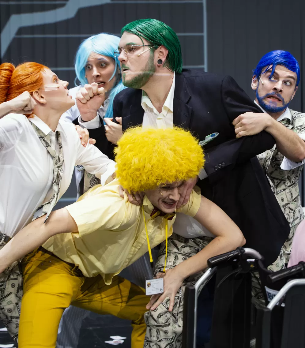 Ensemble mit bunten Perücken und Kostümen, teilweise mit Geldscheinen bedruckt, geraten in eine Prügelei. 