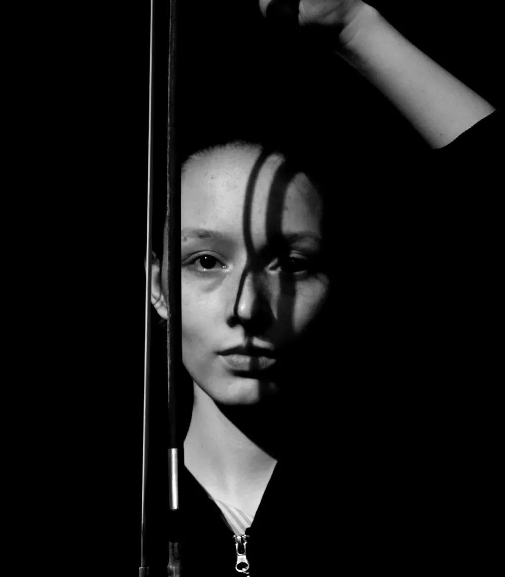 Schwarz-Weiß-Nahaufnahme einer jungen Frau, die einen Cello-Bogen hält, der einen Schatten auf ihr Gesicht wirft