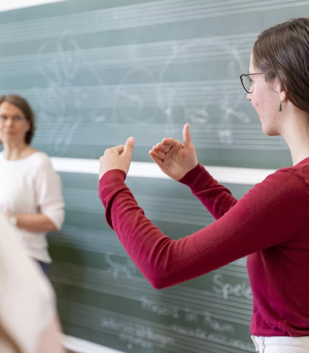 Eine Studentin im roten Pulli steht vor einer Tafel mit Notenlinien und erläutert etwas, dabei gestikuliert sie mit den Händen.