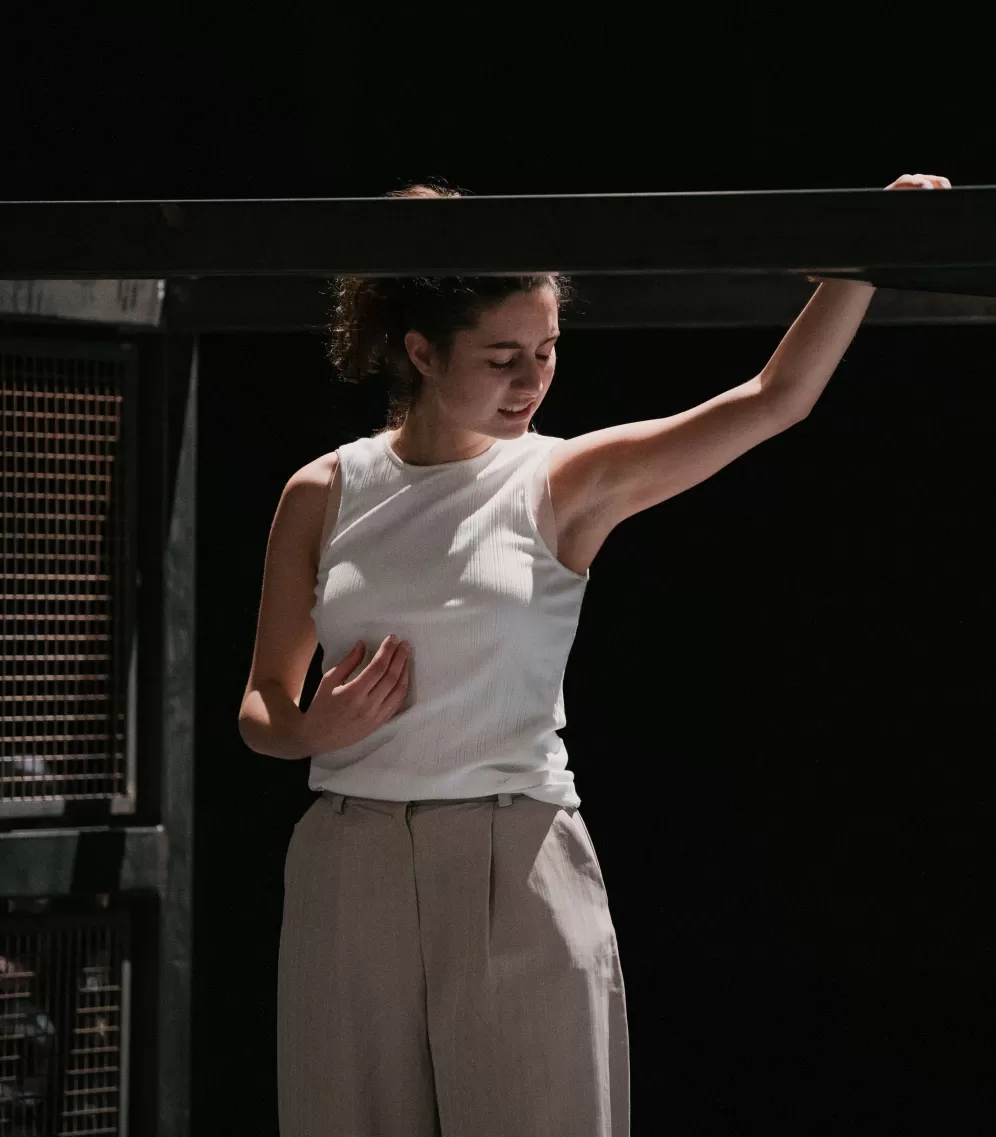 Theaterszene, Darstellerin steht vor Gitterkäfig, ihr Gesichtsausdruck ist schmerzverzehrt.