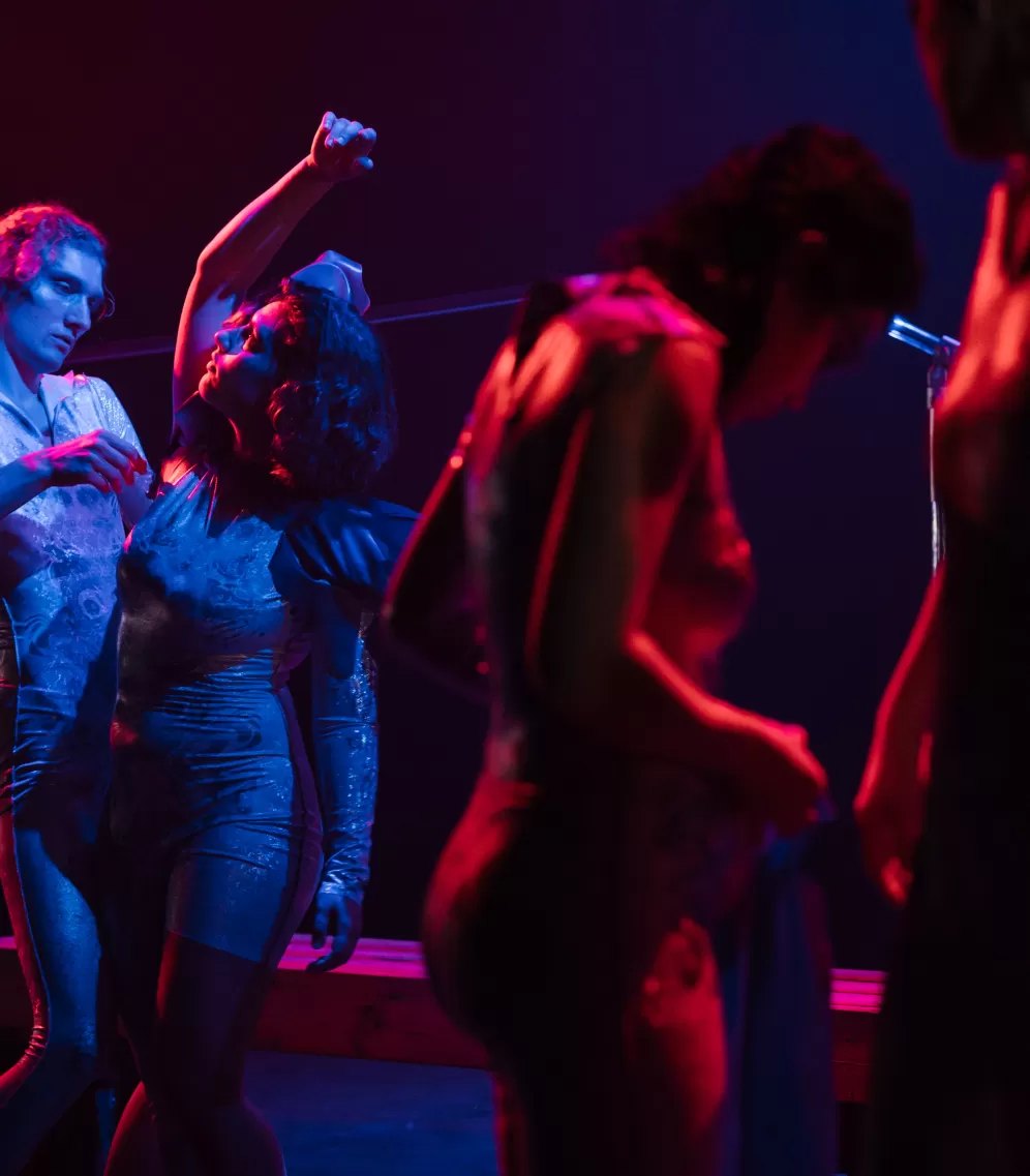 Theaterszene mit tanzenden Menschen im rot-blauen Licht, links zwei Personen im hellen Fokus.