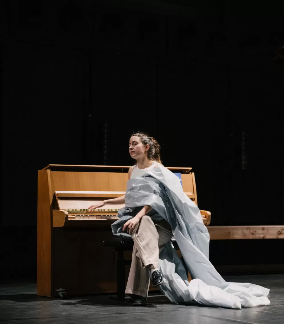 Theaterszene, eine Darstellerin sitzt in einem bauschigen weißen Kostüm an einem Flügel, kehrt den Tasten jedoch den Rücken zu