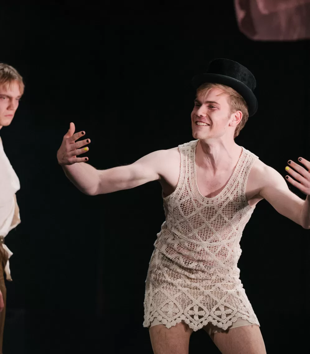 Theaterszene: Zwei Darsteller, einer in Spitzenkleid und Hut, der die Arme ausbreitet.