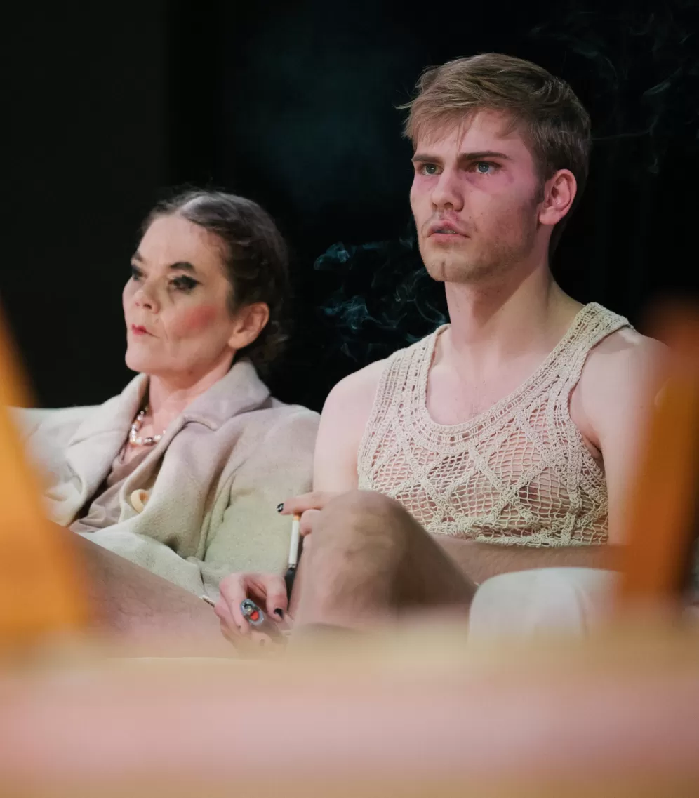 Theaterszene, Nahaufnahme von zwei Darsteller*innen, die nebeneinander sitzen und missmutig aussehen.