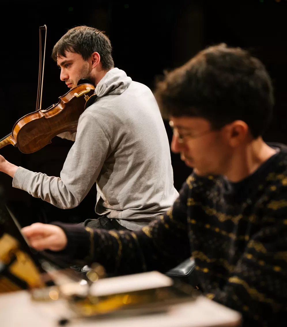 Ein Geiger und ein Pianist während einer gemeinsamen Kammermusikprobe, der Bildfokus liegt auf dem Geiger im Hintergrund des Bildes.