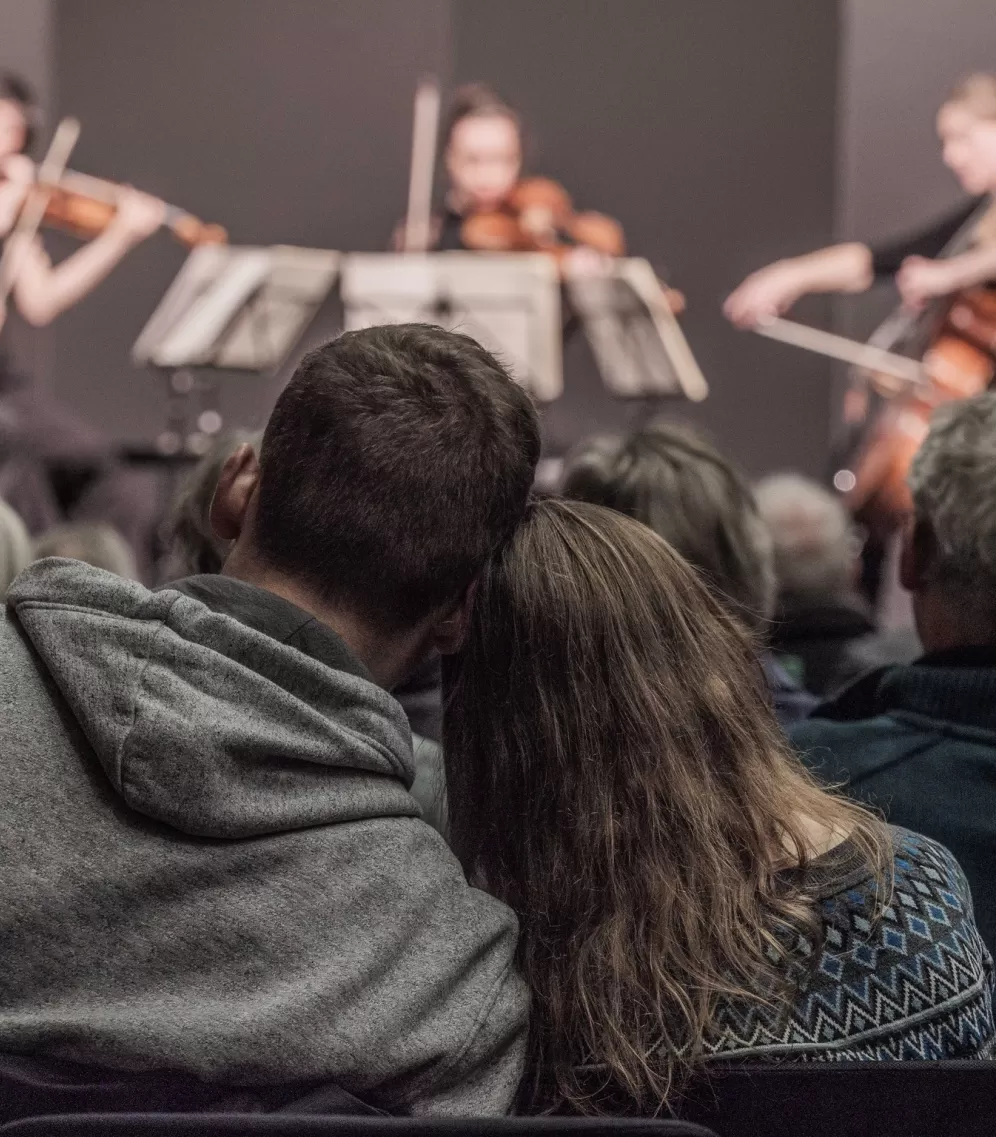 Zwei sitzende Personen lehnen ihre Köpfe aneinander. Im Hintergrund eine Bühne mit einem Streichquartett.