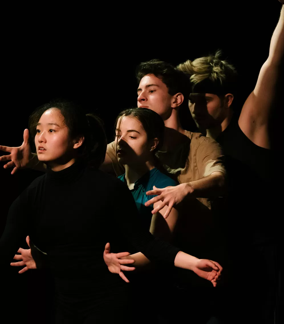 Vier Tänzer*innen stehen hintereinander, sie sind dunkel gekleidet und strecken ihre Arme in unterschiedliche Richtungen aus.