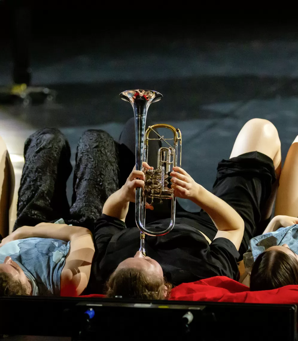 Fünf Personen liegen auf der Bühne auf dem Boden. Die Person in der Mitte spielt Trompete.