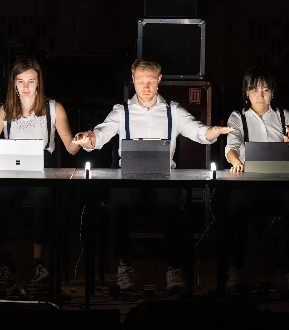 Drei Performer*innen im dunklen Raum, vor ihnen Laptops auf dem Tisch.