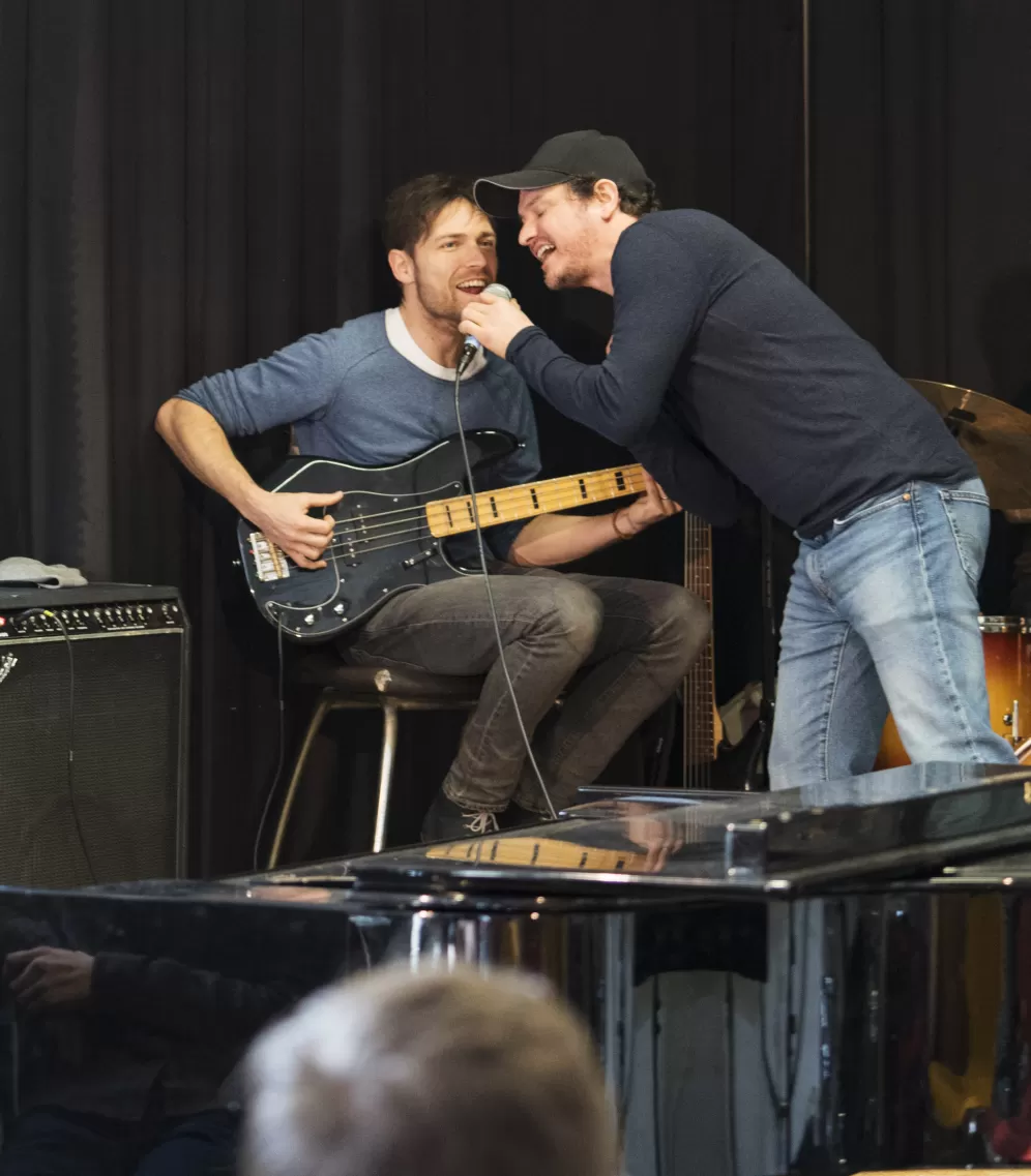 Ein interner Auftritt der Coach-Band. Zwei Coaches singen zusammen in ein Mikrofon. Einer von beiden sitzt auf einem Stuhl und spielt E-Bass.