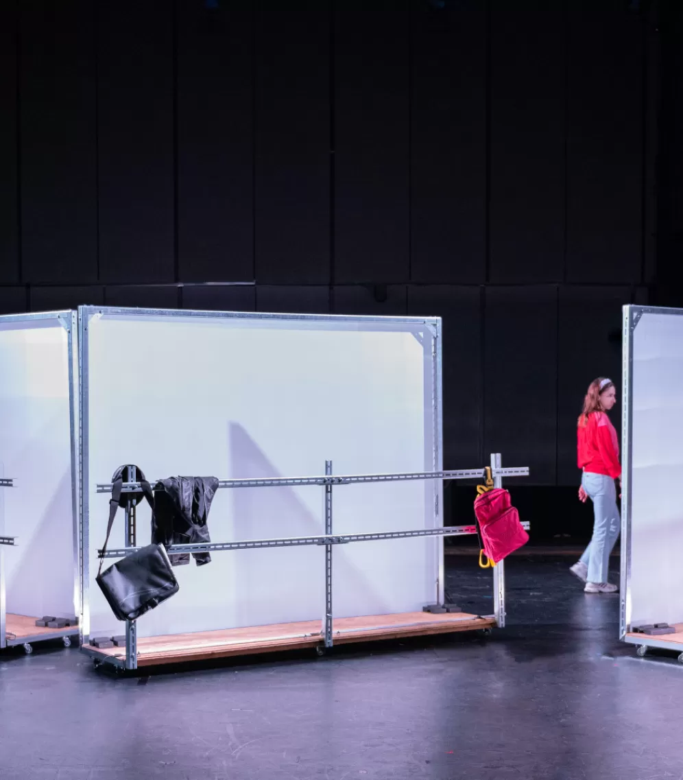 Drei einzelne Stellwände, weiß und leicht transparent, mit jeweils einer Metallbrüstung davor sind schräg gegeneinaner angeordnet, am linken Rand ist ein Mann zu sehen, in der Mitte eine Frau. An den Metallbrütungen hängen Kleidungsstücke und Taschen.