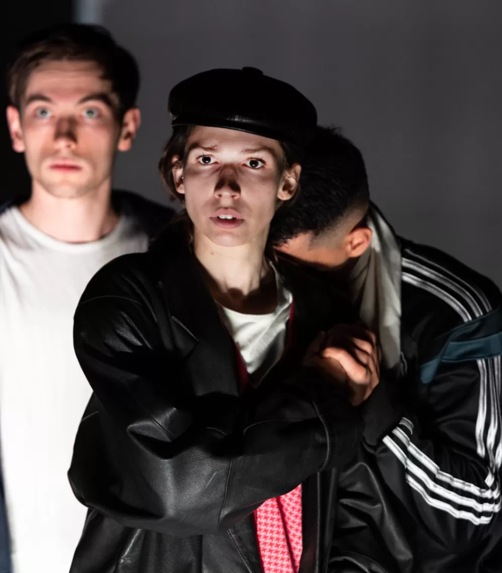 Hintereinander stehend sind drei Personen zu sehen, im Anschnitt bis zur Hüfte. Vorn steht eine Frau in einer schwarzen Lederjacke und Lederkappe auf dem Kopf, an sie lehnt sich seitlich rechts hinten ein Mann, er verbirgt seinen Kopf an ihrer Schulter. Links im Hintergrund ein zweiter Mann, der nach vorn starrt.