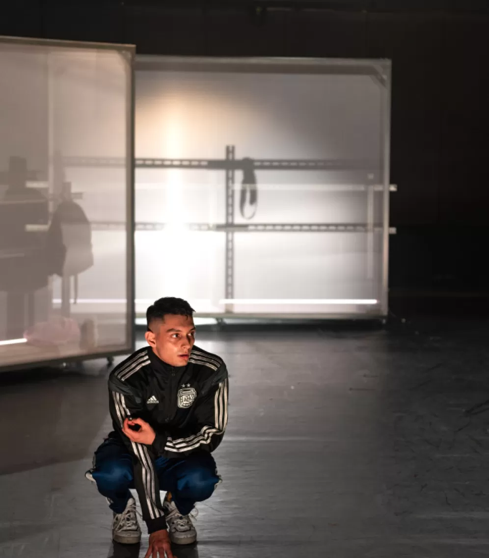 Ein Ausschnitt eines Bühnenraumes, im Hintergrund zwei weiße, halbtransparente Stellwände mit Metallbrüstung davor. Im Vordergrund hockt ein Mann in Turnschuhen und Adidas Trainingsanzug, er schaut fragend nach rechts.