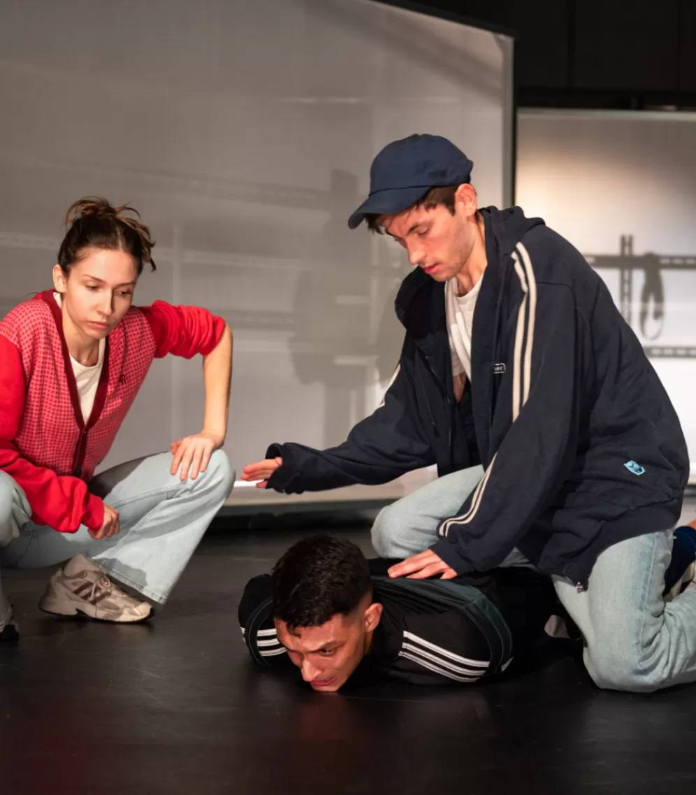Auf dem Bild sind drei Personen in einem Bühnenraum zu sehen. Rechts zwei Männer, einer davon liegt bäuchlings am Boden, der andere kniet mit einem Knie auf ihm und drückt ihn nieder. Links daneben hockt eine Frau in Jeans, Turnschuhen und roter Jacke, die auf den am Boden liegenden Mann blickt.