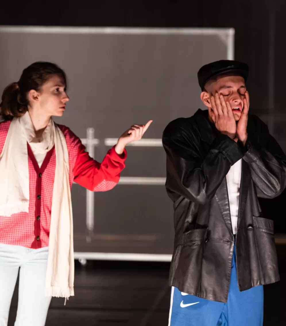 Zwei Personen stehen nebeneinander, sie sind zu sehen bis zum Knie. Links eine Frau mit hellem Schal, weißer Hose und roter Jacke. Sie hat die linke Hand gehoben und zeigt mit dem Zeigefinger auf einen Mann, der rechts neben ihr steht. Der Mann trägt eine schwarze Lederjacke und blaue Nike Sporthose, er hat die Hände seitlich an seine Wangen geschlagen und blickt verzweifelt.