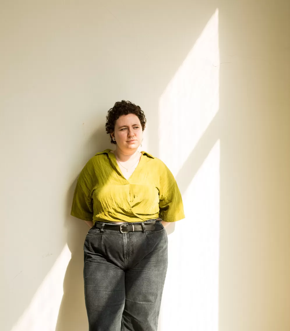 eine Frau mit kurzen braunen Locken steht an einer Wand gelehnt. Sie trägt ein grünes Top und graue Jeans. Die Sonne scheint in ihr Gesicht.