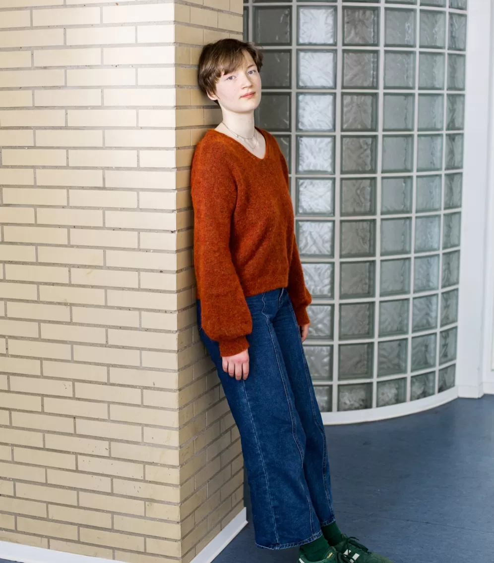 Lilith an der Wand lehnend im roten Pullover. 