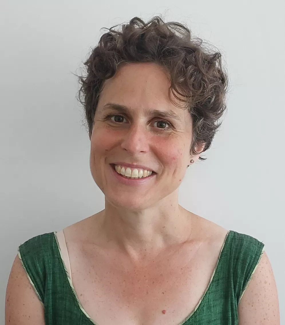 Katrin Raetzel hat kurze braune Locken, braune Augen und trägt ein grünes T-Shirt. Sie steht vor einer weißen Wand und lächelt in die Kamera.
