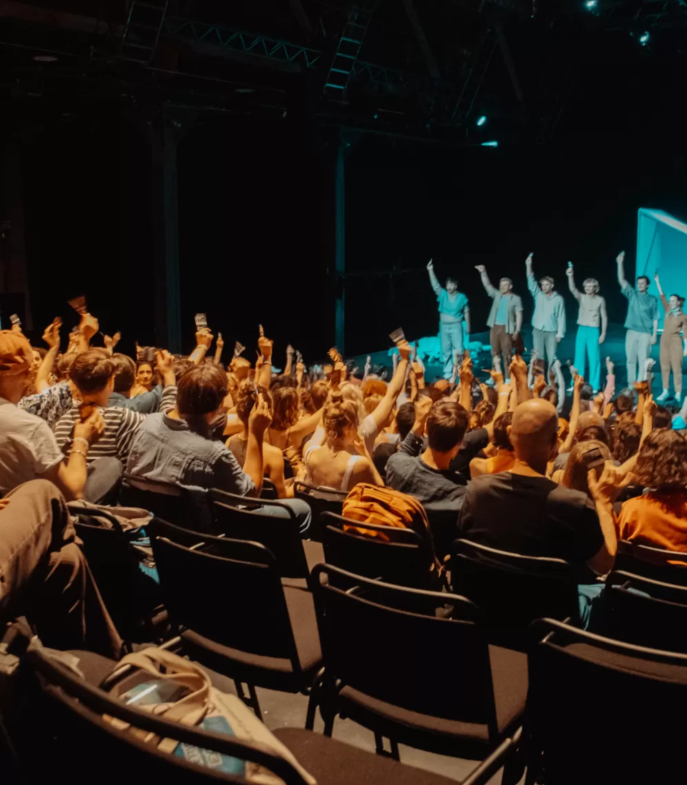 Publikum im Bockenheimer Depot, Blick auf die Bühne wo eine Gruppe Darsteller*innen das Publikum zum Mitmachen motiviert.
