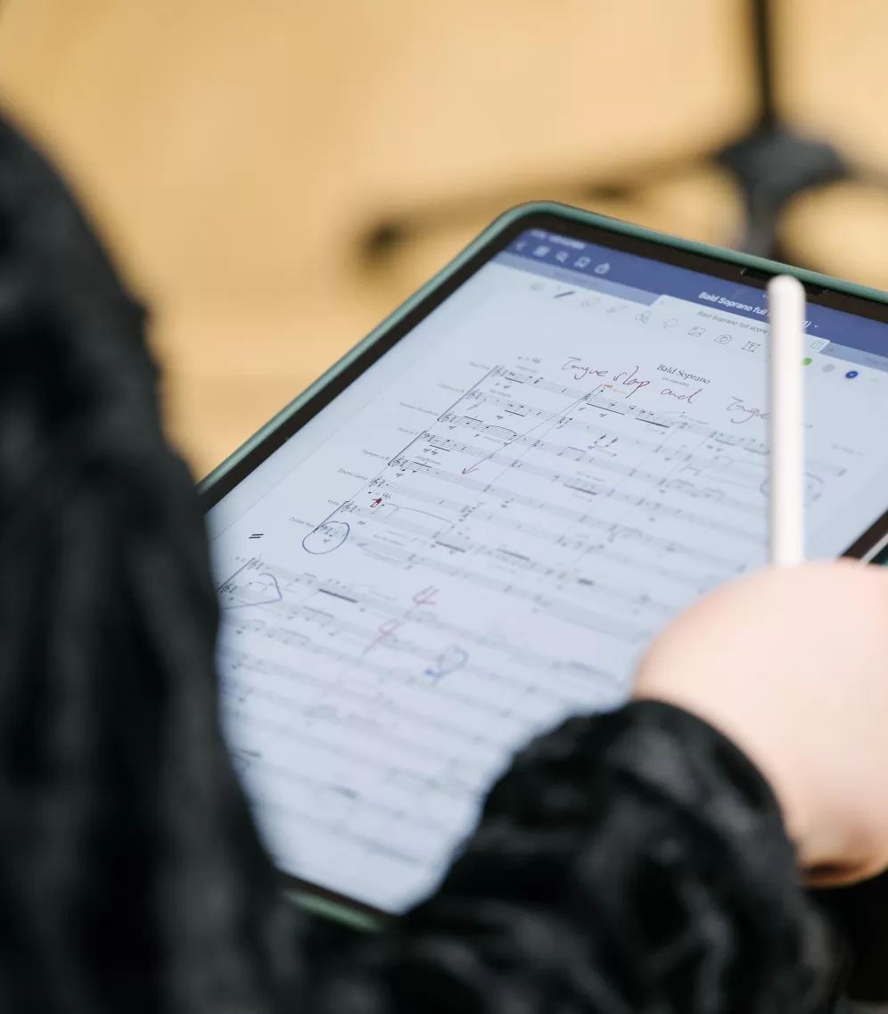 Nahaufnahme eines Tablet-Computers auf den Knien einer Person, die einen Stift in der Hand hält. Auf dem Display sind Noten zu sehen.