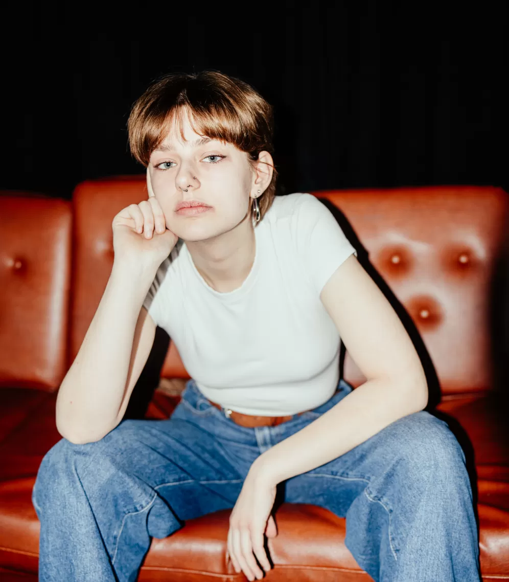 Halbtotale Paula Marie Schulze, weißes T-Shirt, blaue Jeans, im Hintergrund eine braune Ledercouch