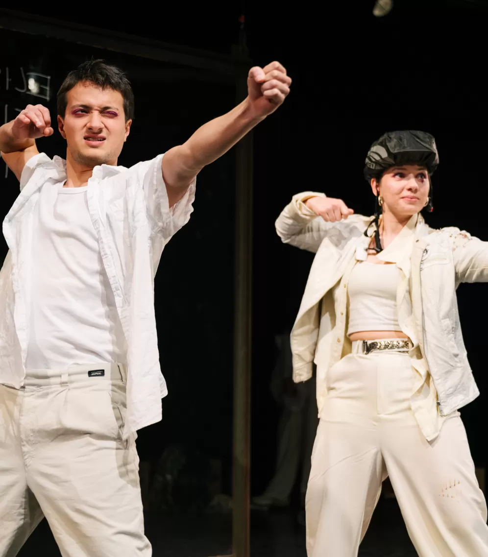 Zwei ganz in weiß gekleidete Schauspieler*innen, die so tun als ob sie Pfeil und Bogen abschießen.