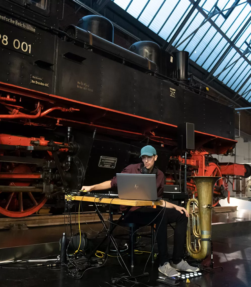 Ein Mann sitzt an einem Tisch und schaut auf seinen Laptop. Hinter ihm steht eine riesige Dampflock, neben ihm eine Tuba.