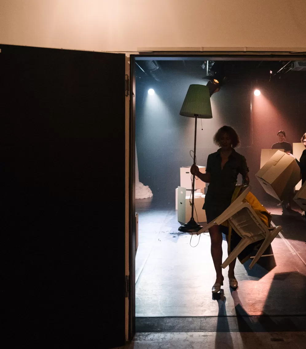 Eine Person mit Stehlampe und Umzugskarton in der Hand kommt aus einem Theatersaal.