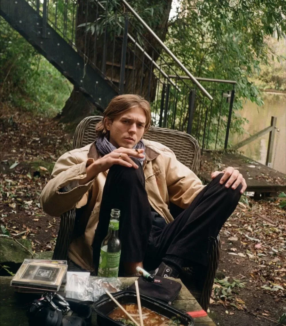 Ein junger, hübscher  Mann sitzt rasuchend an einem Tisch, am Ufer eines Flusses. Auf dem Tisch liegen CDs und Essen.