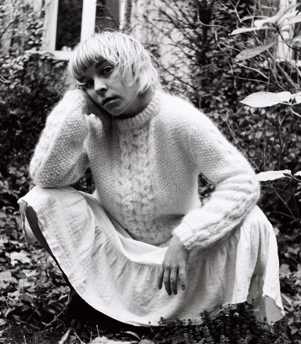 Ein schwarz-weiß Bild. Man sieht eine Person in der Hocke. Ringsherum Efeu und andere Pflanzen. 