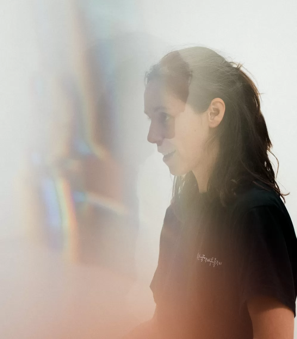 Eine junge Frau, Melissa Ugolini, ist im Profil von der Seite zu sehen. Sie trägt ein dunkles T-Shirt und hat die Haare nach hinten gebunden. Ihr Gesicht ist leicht beleuchtet, während weiche, bunte Lichtreflexe und ein diffuser Schleier im Vordergrund das Bild teilweise überlagern. Der Hintergrund ist schlicht und hell, wodurch die Person ruhig und nachdenklich wirkt.