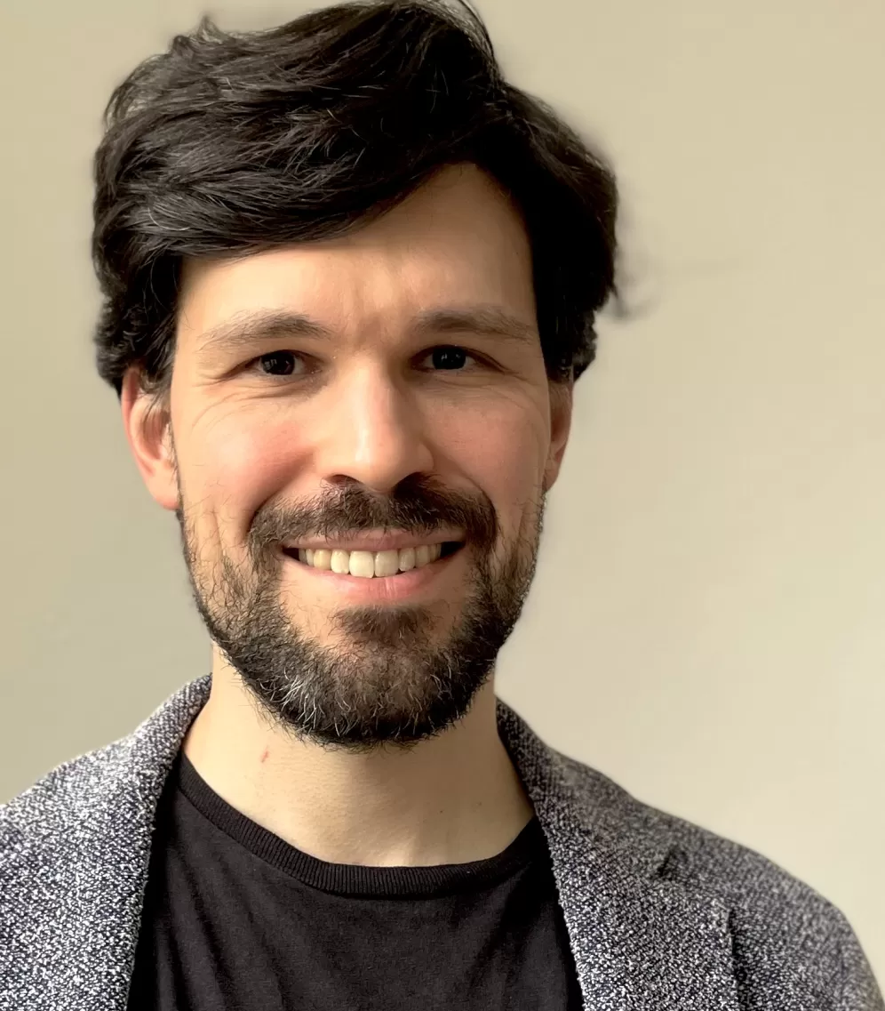 Ein Porträtfoto von Diego Ramos Rodríguez, Mann, mitte dreißig, dunkle Haare und Bart, lächelnd zur Kamera, schwarzem T-Shirt und grauer Jacke